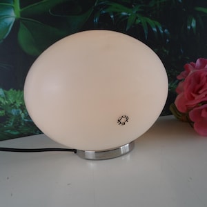 Op de afbeelding: Een bolvormige, crèmekleurige tafellamp met een chromen voet. De lamp heeft een klein, zwart logo op het oppervlak. Een boeket roze rozen staat rechts van de lamp. De lamp is aangesloten op een zwart netsnoer.