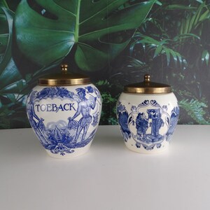 Pode incluir: Dois vasos de cerâmica azul e branca com tampas douradas. O vaso maior tem a palavra "TOEBACk" escrita nele. Ambos os vasos têm um design de inspiração holandesa.