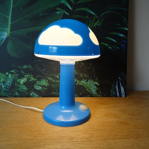 Peut inclure: Une lampe de table en forme de champignon bleue avec un motif de nuage blanc sur le dessus. La lampe a un cordon blanc et un bouton blanc sur la base.