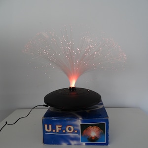 Op de afbeelding: Een zwarte UFO-vormige glasvezellamp met een witte glasvezelpluim die van boven uitstraalt. De lamp is aangesloten en brandt, met roze licht dat door de vezels schijnt. De lamp staat op een blauwe doos met de tekst "U.F.O." erop.