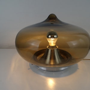 Può includere: Una lampada da tavolo moderna di metà secolo con un paralume in vetro marrone e una base argentata. La lampada è accesa con una lampadina.