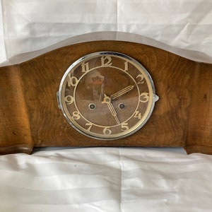 Puede incluir: Un reloj de chimenea de madera vintage con una esfera redonda con borde plateado y marcadores de hora romanos negros. El reloj tiene un acabado de madera marrón claro y una forma ligeramente arqueada.