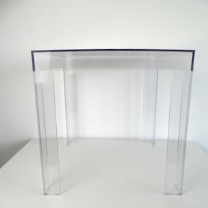 Può includere: Un tavolino laterale in acrilico trasparente con un piano rettangolare e quattro gambe. Il tavolo è trasparente, permettendo di vedere lo sfondo attraverso di esso. Il tavolo è alto circa 46 cm.