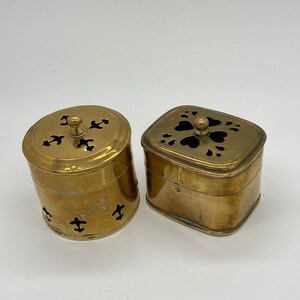 Op de afbeelding: Twee messing containers met deksels. De containers hebben ingewikkelde uitgesneden ontwerpen. Een container is rond en de andere is vierkant.