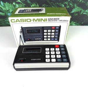 May include: A vintage Casio-Mini CM-603 electronic calculator, displayed with its original box. The calculator is black with white and gray keys, and a red 'C' button. The box is white and green with the calculator's image and text.
