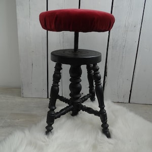 Pode incluir: Um banquinho de madeira preto ajustável em altura com um assento de veludo vermelho. O banquinho tem um design de pernas torneadas e está sentado em um tapete branco e felpudo.
