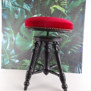 Pode incluir: Um banco vintage de madeira preta com um assento redondo de veludo vermelho. O assento é adornado com pequenos pregos dourados. O banco tem três pernas e um design torneado decorativo. É um móvel clássico.