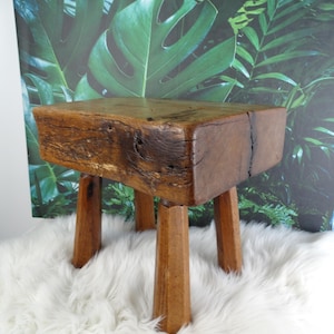 May include: A small, wooden stool with a dark brown, weathered finish. It has a rectangular top and four angled legs. The stool is placed on a fluffy, white rug, with a backdrop of green tropical foliage. The stool's dimensions are approximately 30 cm x 30 cm x 30 cm.