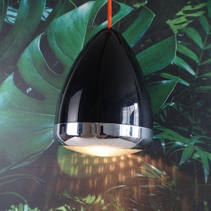 May include: A black, teardrop-shaped pendant light with a chrome band and an orange cord. The light is suspended against a backdrop of green tropical foliage. The light is turned on, casting a warm glow.