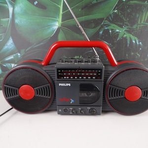 Könnte beinhalten: Ein schwarzer und roter Kassettenspieler mit einem roten Griff und zwei großen Lautsprechern. Der Spieler hat eine silberne Antenne und den Text "PHILIPS" und "Radio-Recorder" auf der Vorderseite.