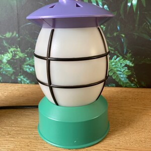 May include: A white and green table lamp with a purple top and a black wire cage. The lamp has a round base and a round top with a small hole in the centre.