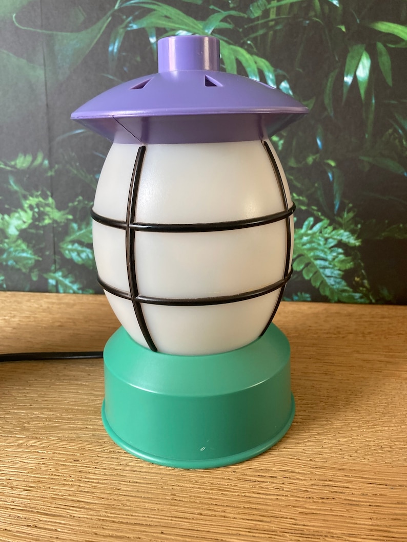May include: A white and green table lamp with a purple top and a black wire cage. The lamp has a round base and a round top with a small hole in the centre.