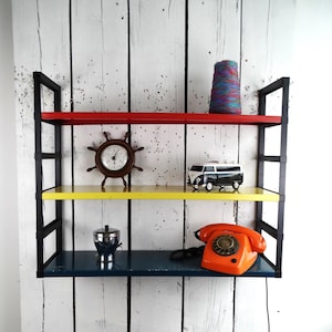 May include: A three-tiered shelf unit with red, yellow, and blue shelves. The shelves hold decorative items, including a clock, a toy van, a silver container, an orange rotary phone, and a spool of yarn. The unit has black metal supports and is mounted on a white wooden background.