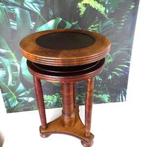 Pode incluir: Uma mesa lateral de madeira vintage com tampo redondo e três pernas. A mesa tem um tampo castanho escuro com uma moldura de madeira castanha mais clara. As pernas são de cor castanha escura e a mesa tem aproximadamente 61 cm de altura.