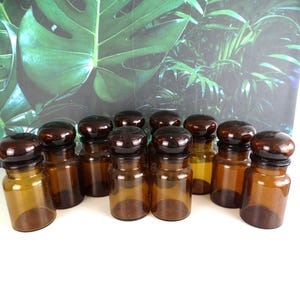 Op de afbeelding: Een verzameling van tien kleine apothekerspotten van amberkleurig glas met bijpassende ronde bruine glazen deksels. Elke pot heeft een cilindrisch lichaam en een korte hals, gerangschikt in een groep om het bruine glas te tonen.