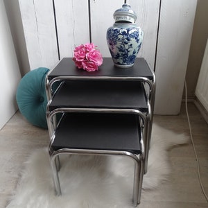 Puede incluir: Un conjunto de tres mesas nido negras con patas cromadas. Las mesas están sobre una alfombra blanca y esponjosa. Un jarrón de porcelana azul y blanco con tapa se encuentra en la mesa superior, y un ramo de rosas rosas se encuentra en la mesa del medio.