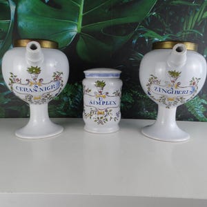 Op de afbeelding: Drie antieke apothekerspotten van keramiek met bloemmotieven en gouden accenten. De potten zijn gelabeld met "CERAS.NIGR", "U. SIMPLEX" en "ZINGIBERIS".