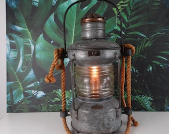 Op de afbeelding: Een vintage-stijl metalen lantaarn met een touwhandvat en een heldere glazen bol. De lantaarn is verlicht en heeft een warme gloed.