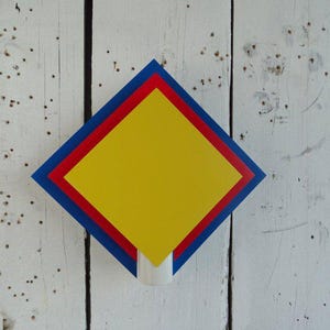 May include: A geometric wall art piece featuring a yellow square within red and blue borders. The artwork is mounted on a white wooden surface. The design is simple and modern, with a pop of color.