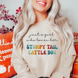 Può includere: Una felpa beige con il testo "just a girl who loves her STUMPY TAIL CATTLE DOG" in colori arcobaleno. La felpa è indossata da una persona seduta su un divano.