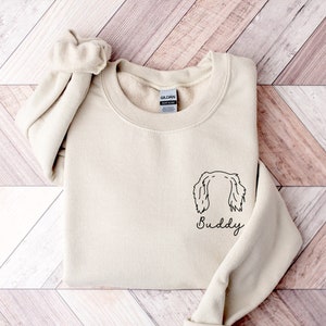 May include: A beige sweatshirt with a black line drawing of a dog's head and the word "Buddy" underneath.