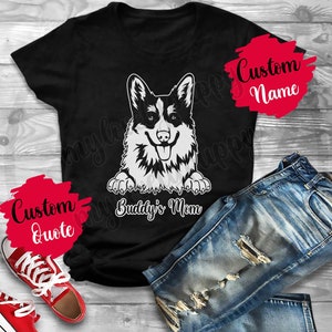 May include: Black t-shirt with a white graphic of a corgi dog peeking over the top. The text "Buddy's Mom" is printed below the dog. The shirt is laying on a wooden surface with a pair of blue jeans and red sneakers.