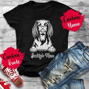 Pode incluir: Camiseta preta com um gráfico branco de um cão dachshund com o texto "Buddy's Mom" embaixo. A camiseta está sobre uma superfície de madeira com um par de calças jeans azuis e tênis vermelhos.