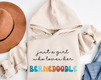 Può includere: Una felpa con cappuccio beige con il testo "just a girl who loves her BERNEDoodle" in lettere nere. La parola "BERNEDoodle" è in lettere colorate e stilizzate.