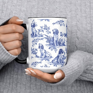 Op de afbeelding: Witte keramische mok met een zwart handvat en rand. De mok heeft een blauw en wit toile-patroon van honden in verschillende poses te midden van gebladerte. De mok wordt vastgehouden door een persoon die een grijze trui draagt.