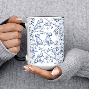 May include: White ceramic mug with a black handle and rim, featuring a blue and white toile pattern of dogs and floral elements. The mug is held by a person wearing a gray sweater.
