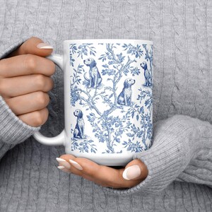May include: White ceramic mug with a blue and white toile pattern depicting dogs and floral motifs. The mug has a large handle and a cylindrical shape. The design is repeated around the mug.