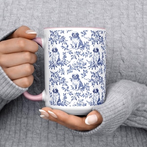 Pode incluir: Caneca de cerâmica branca com um padrão de cão e floral azul e branco. A caneca tem interior e alça rosa. O design apresenta múltiplas ilustrações de cães em meio a um padrão de folhas e videiras. Um item decorativo.