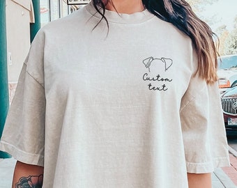 May include: A light beige oversized t-shirt with a simple line drawing of a dog with the text "Custom text" underneath. The person wearing the shirt is standing on a sidewalk in front of a tree.