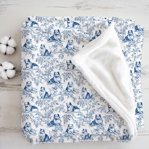 Puede incluir: Una manta blanca y azul con un patrón repetido de perros y follaje. Los perros están representados en un estilo toile clásico, con un reverso de felpa blanco y suave. La manta está doblada, revelando el reverso de felpa.