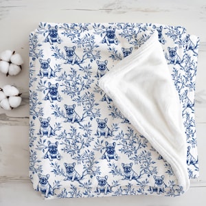 May include: A white baby blanket with a blue French bulldog and floral pattern. The blanket has a soft, white, plush backing. The blanket is folded, revealing the plush backing. Two cotton buds are in the upper left corner.