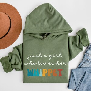 Może przedstawiać: Zielony sweter z kapturem z napisem "just a girl who loves her WHIPPET" w białych i kolorowych literach. Sweter leży płasko na białym tle z brązowym kapeluszem i niebieskimi dżinsami.