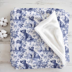 May include: A white and blue blanket with a repeating pattern of Shih Tzu dogs and floral elements. The blanket has a soft, white, plush underside. The design is reminiscent of classic toile patterns.