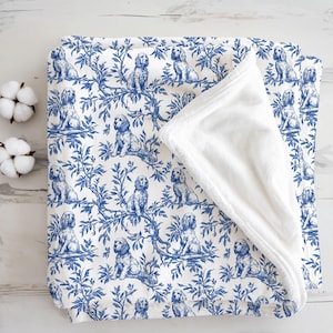 Può includere: Una coperta bianca e blu con un motivo ripetuto di cani sui rami. La coperta ha un lato inferiore in morbido peluche bianco. Il design ricorda i classici motivi toile.