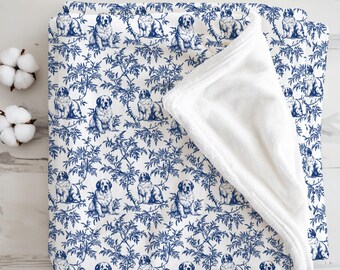 May include: A white and blue blanket with a repeating pattern of seated dogs and foliage. The blanket has a soft, white, plush underside. The design is reminiscent of toile fabric. Two cotton buds are visible in the upper left corner.
