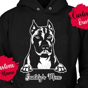 Pode incluir: Camisola de capuz preta com uma silhueta branca de um pit bull a espreitar por cima. O texto "Buddy's Mom" está impresso abaixo do cão. A camisola tem uma etiqueta de nome personalizada vermelha na manga esquerda e uma etiqueta de citação personalizada vermelha na manga direita.