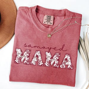 May include: A dusty rose t-shirt with the words "samoyed MAMA" in a floral design. The shirt has a crew neck and short sleeves. A gold necklace with a small pendant rests on the shirt. A brown hat and dried leaves are in the background.