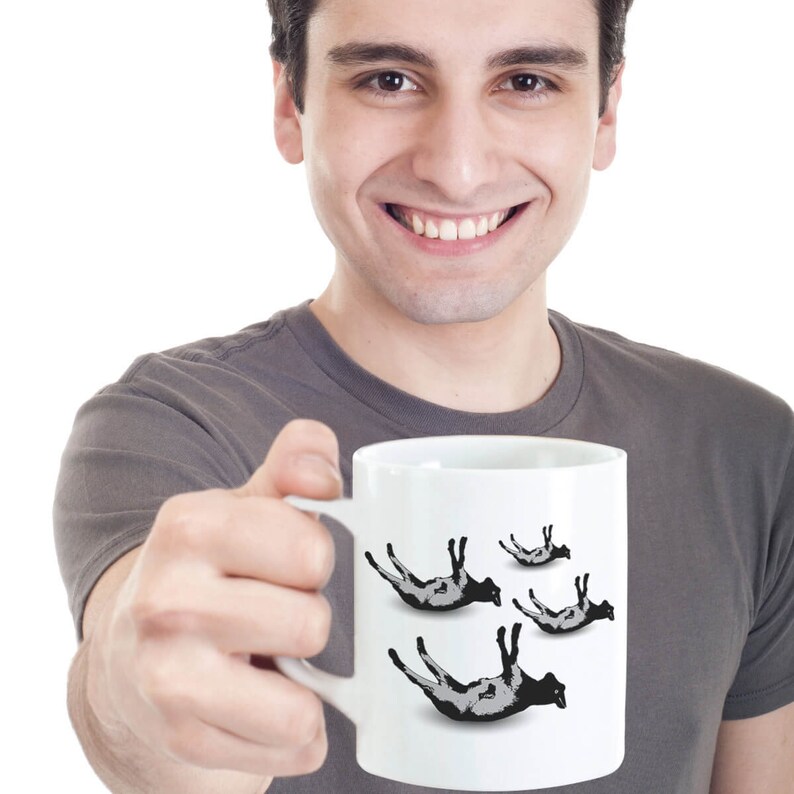 May include: A white ceramic mug with a black and white graphic of sheep falling from the sky. The mug is being held by a person.