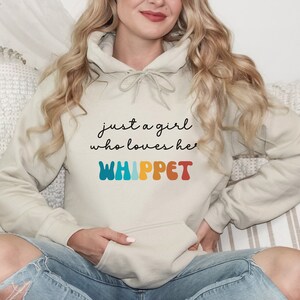 Może przedstawiać: Jasnobeżowa bluza z kapturem z napisem "just a girl who loves her WHIPPET" w kolorowej czcionce retro.
