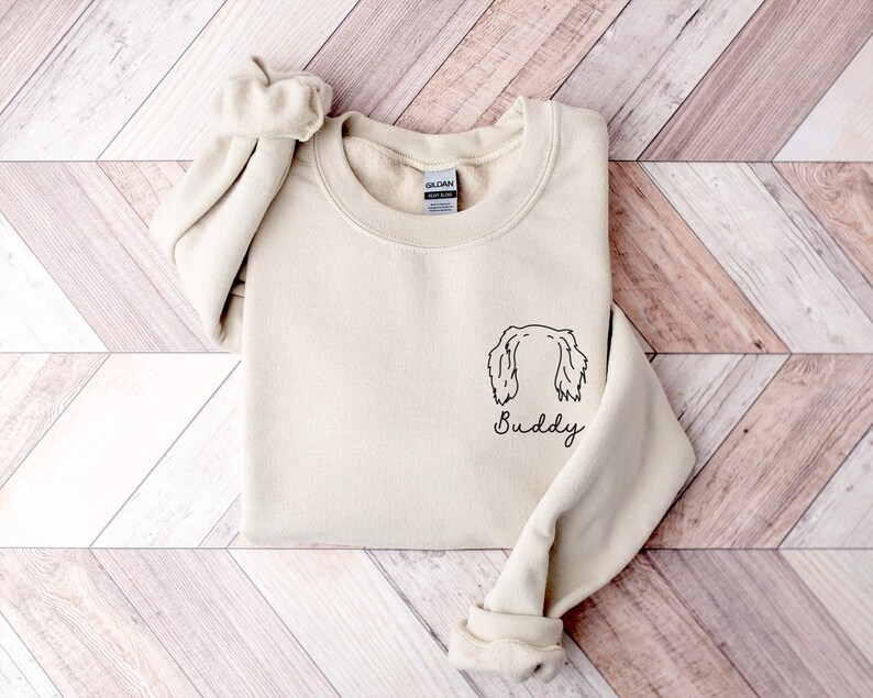May include: A beige sweatshirt with a black line drawing of a dog's head and the word "Buddy" underneath.
