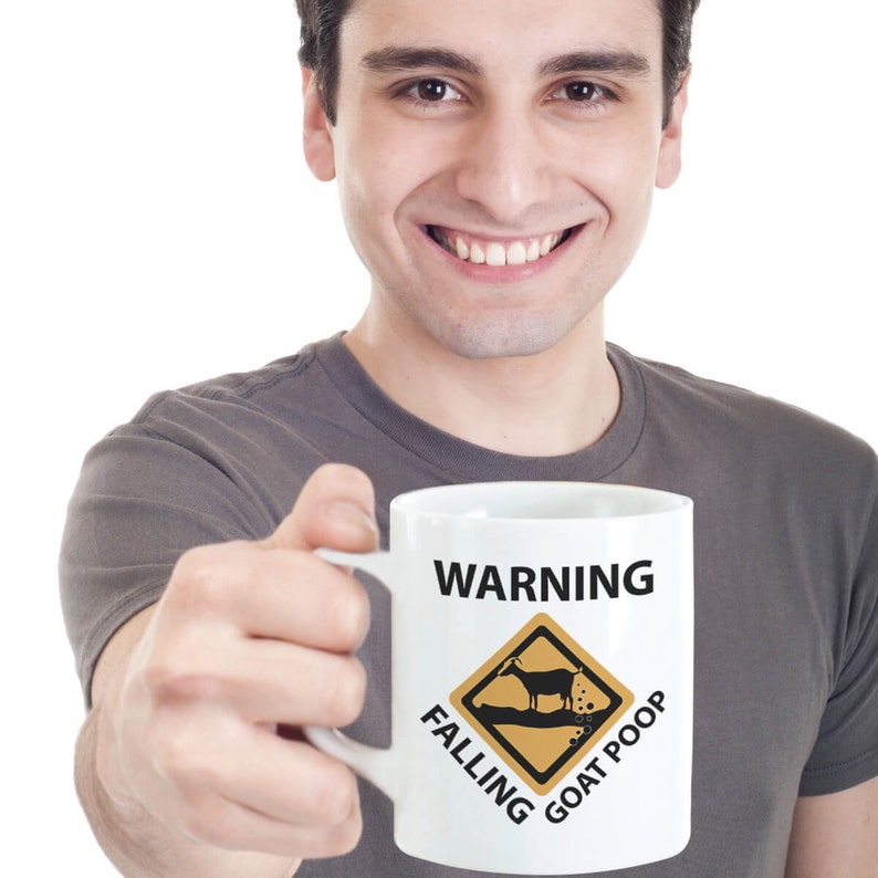 Puede incluir: Una taza de caf&eacute; blanca con un letrero de advertencia negro que dice "WARNING FALLING GOAT POOP". El letrero es un diamante amarillo con una silueta negra de una cabra que cae y hace caca. Una persona sostiene la taza en su mano derecha.