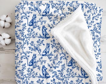 May include: A white and blue blanket featuring a repeating pattern of dogs and foliage. The blanket has a soft, white, plush underside. Two cotton buds are visible in the upper left corner. The blanket is folded on a light wood surface.