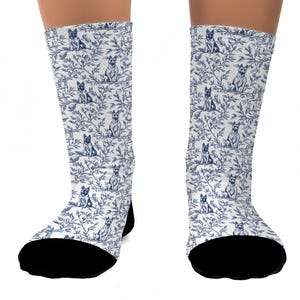 May include: A pair of knee-high socks featuring a repeating pattern of blue dogs and foliage on a white background. The socks have black toes and heels. The design resembles a classic toile pattern.