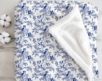 May include: A white and blue blanket with a toile pattern featuring dogs in trees. The blanket has a soft, white, plush backing. Two cotton buds are visible in the corner.