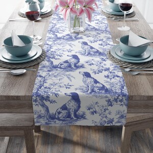 May include: A white table runner featuring a repeating blue toile pattern of dogs sitting among trees and floral branches. The fabric is laid flat across a wooden table, serving as a decorative base for place settings with bowls and glasses.