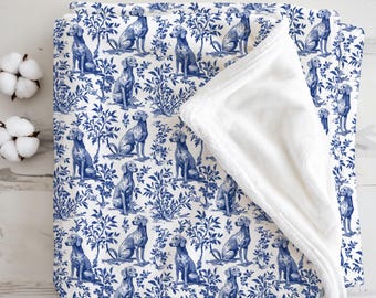 May include: A white and blue patterned blanket with a repeating design of dogs and foliage. The blanket has a soft, white, plush underside. The dogs are depicted in a classic toile style, with a blue and white colour scheme.